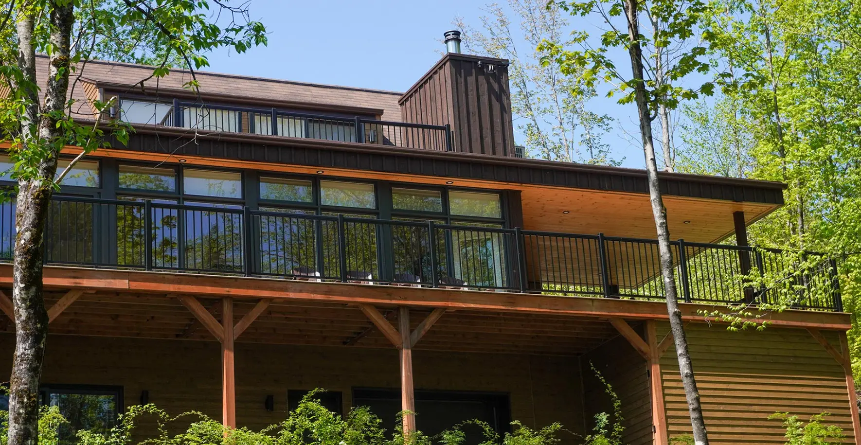 A low-angle view of a modern cottage, surrounded by lush green trees in the late spring.